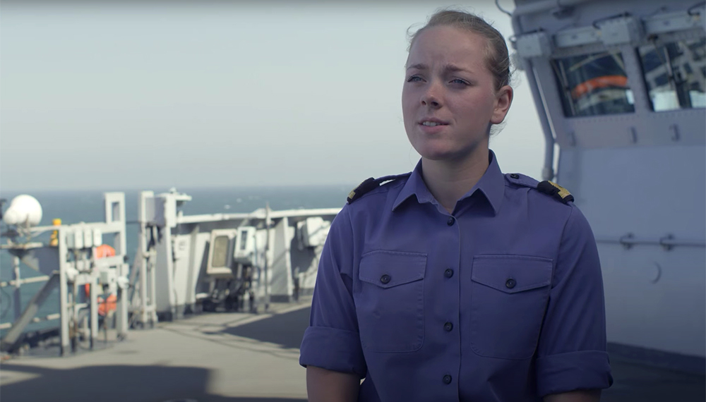 blonde haired blue shirted royal navy crewmember face on with ship bridge and light blue skies in background