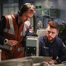 two crewmembers in uniform with one wearing orange safety vest work in onboard operations rooms