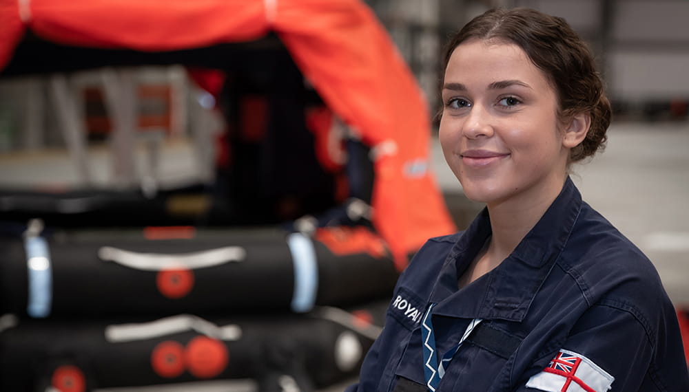 brown-haired smiling blue uniformed Survival equipment technician with black equipment and orange tarpaulin in background