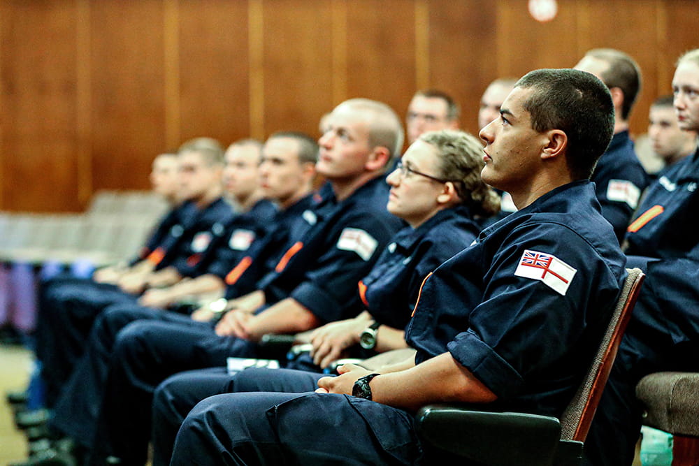 royal navy personnel in blue uniforms sitting down in conference hall looking towards stage on left