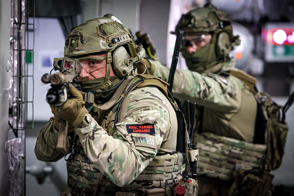 A colour photo shows two Royal Marine Commandos with guns aimed at something just out of frame. They are in an enclosed space and wear full fatigues.