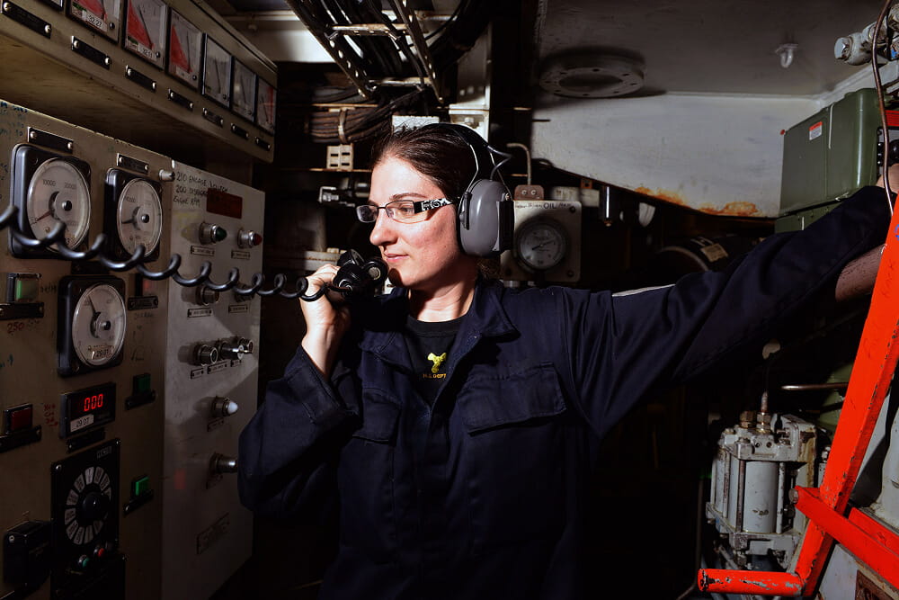 A colour photo shows an RFA engineer in an enclosed space with mechanical equipment. She wears a blue Royal Navy work uniform and protective headset. She speaks into a black telephone handset that connects to the equipment. There are several dials and lights on the panels.
