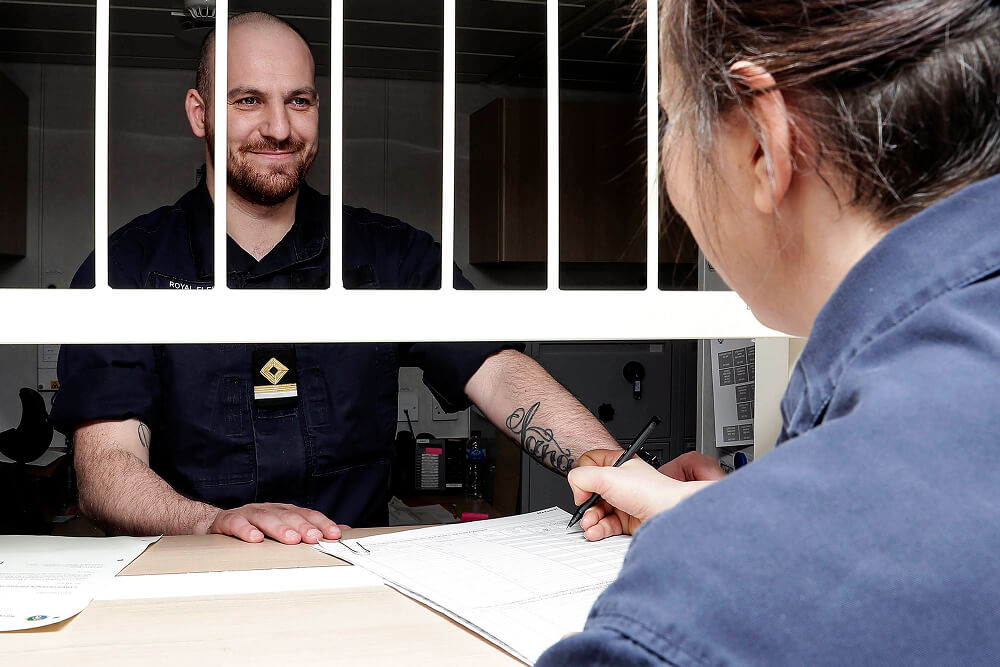 A colour photo shows an over-the-shoulder perspective of an RFA logistics officer standing behind a service counter. He smiles at a servicemember who stands on the other side of the counter; she is filling out a form. They both wear blue Royal Navy work uniforms.