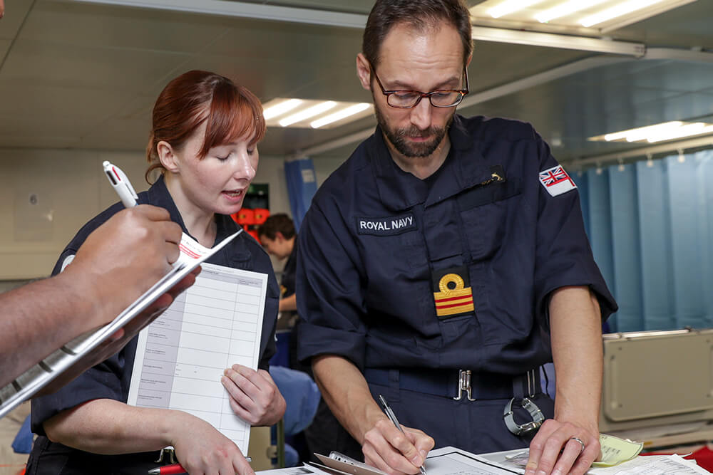 A colour image shows a Submariner physician taking medical notes in an infirmary. Standing next to him, a female Submariner medical professional relays information. They both wear blue Royal Navy work uniforms with Submariner badges. In the foreground, just out of frame, a third medical professional takes notes; the image has been cropped and only a forearm and clipboard is visible. In the background is a glimpse of a fourth medical Submariner in the middle of their duties.