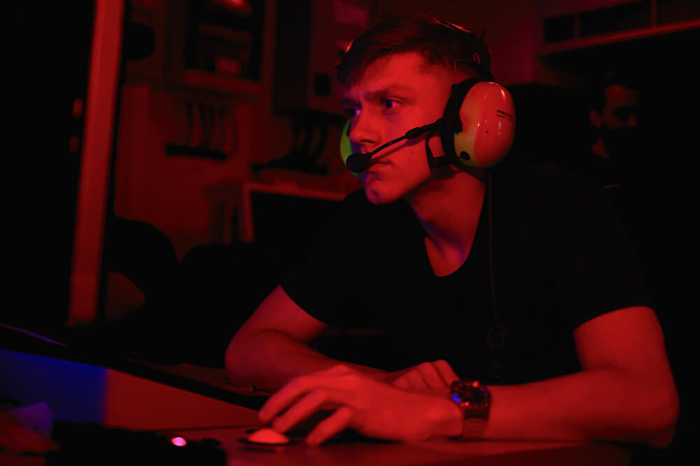 A colour image shows a Submariner engineer sitting at a workstation. He looks intently at equipment just out of frame. He is in a dark room illuminated by red lights. He wears a watch on his wrist and a protective headset.