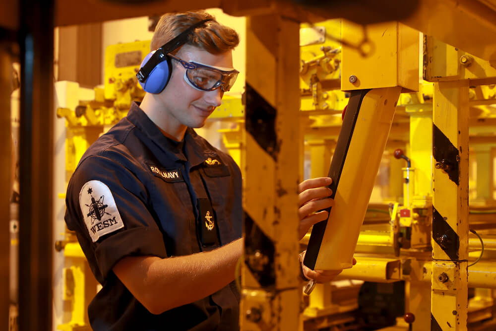 A colour image shows a Submariner engineer examining heavy-duty equipment in an enclosed space. He wears protective earmuffs, protective eye gear, and a blue Royal Navy work uniform with Submariner badges.