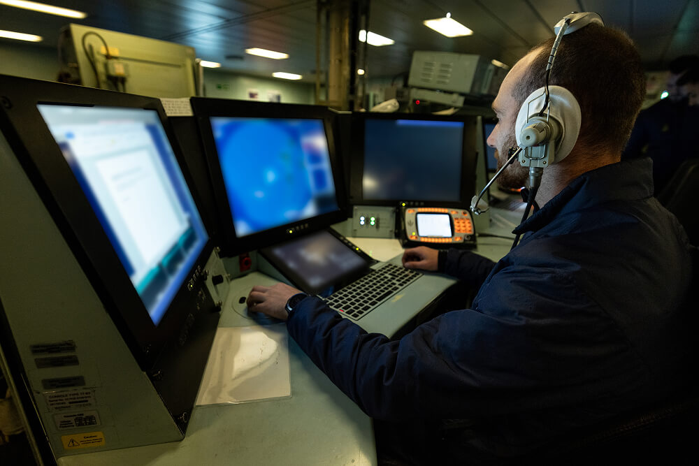 A colour image shows a Royal Navy cyber specialist sitting at a workstation with three large screens, a keyboard, and other desktop tech. He wears a blue Royal Navy work uniform and a protective, heavy-duty headset. In the background are large machines and equipment.