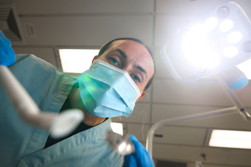 A colour image shows the point-of-view of a dental patient. Looking up from a chair, the patient sees a Royal Navy dental practictioner holding specialist tools. A bright light shines down. The dental practicioner wears light blue scrubs, sterile gloves, and a protective face mask.