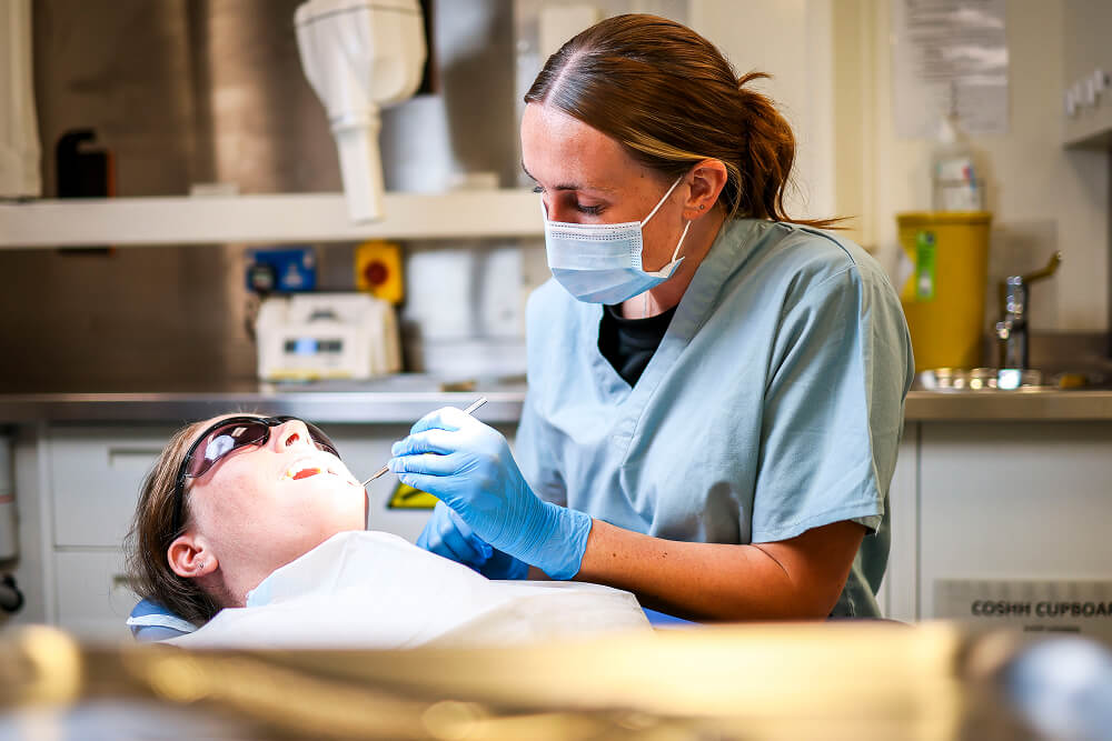 A colour image shows a Royal Navy dental nurse examining a patient's mouth with specialist tools. She wears light blue scrubs, sterile gloves, and a protective face mask. A bright light shines on the patient who wears protective glasses.
