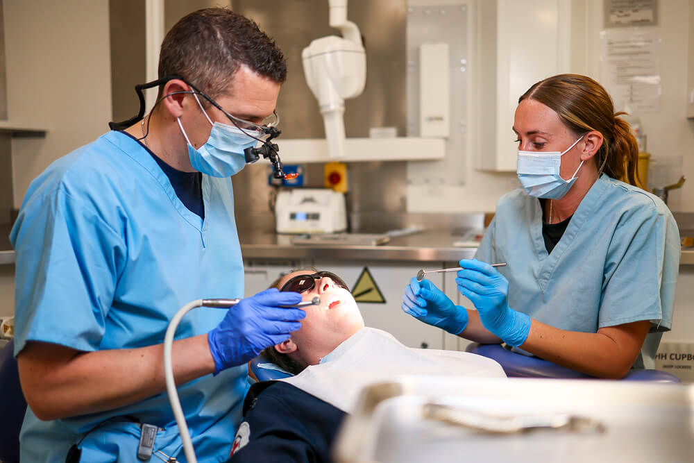 A colour photo shows a Royal Navy dentist and dental assistant performing a procedure on a patient. The patient is laid back and wears protective goggles. A bright light shines on their face. The dentist holds a dental drill in hand and wears magnifying dentist loupes. The assistant holds a dental mirror in hand. Both wear light blue scrubs and sterile gloves.