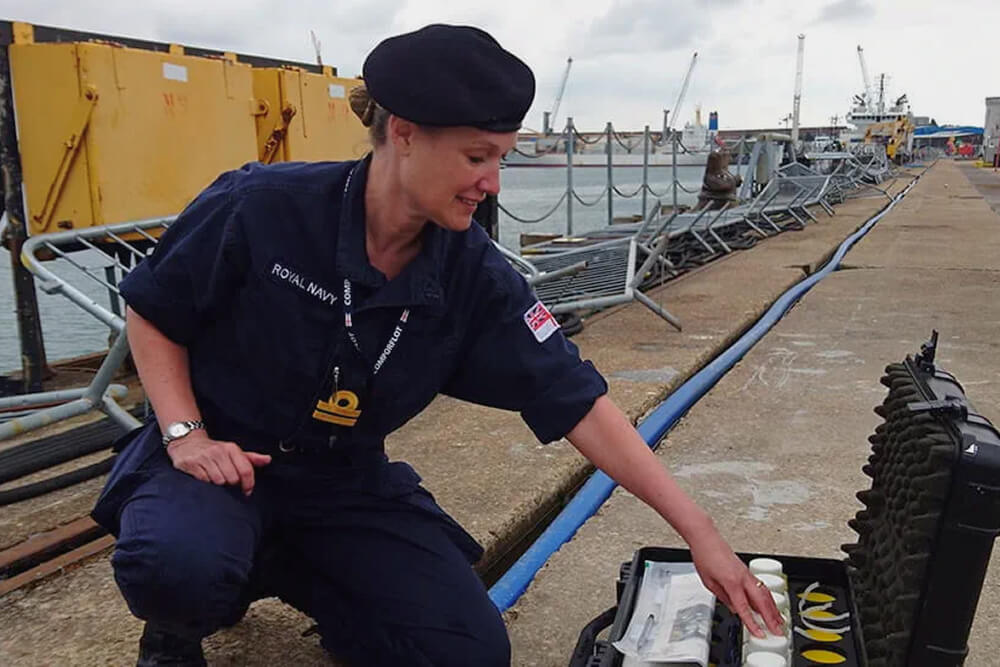 A colour image shows a Royal Navy environmental health officer at an industrial port. She is kneeling down on the concrete and examines the contents of an open kit. Inside are several unidentified bottles and other equipment. She is wearing a blue Royal Navy work uniform, navy blue beret, and silver wrist watch. In the background are several cranes, protective barriers, a glimpse of the ocean, and a bright blue cable that runs the length of the port.