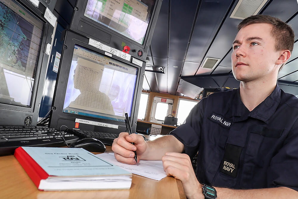 A colour image shows a Royal Navy meteorology specialist sitting at a workstation. He takes notes while observing several digital screens with geographic weather information. He wears a blue Royal Navy work uniform.