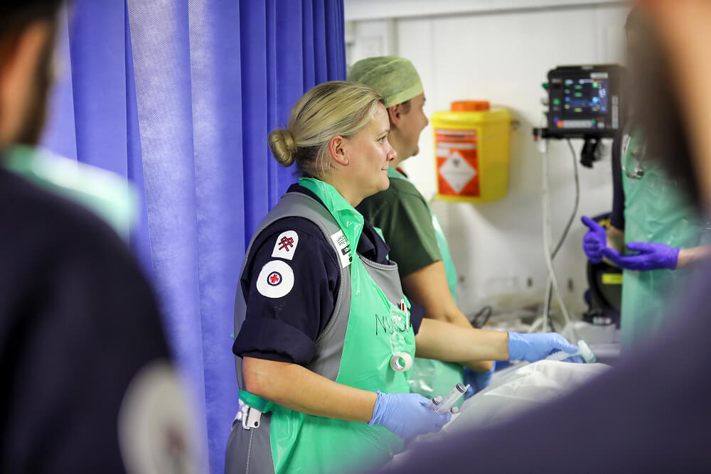 A colour image shows an over-the-shoulder perspective of a Royal Navy medical assistant near a patient's bedside. The bed and patient are  just out of frame. The medical assistant is wearing a blue Royal Navy work uniform, sterile gloves, and a sterile protective vest. In her hand is a syringe. In the background are two more medical personnel in conversation.
