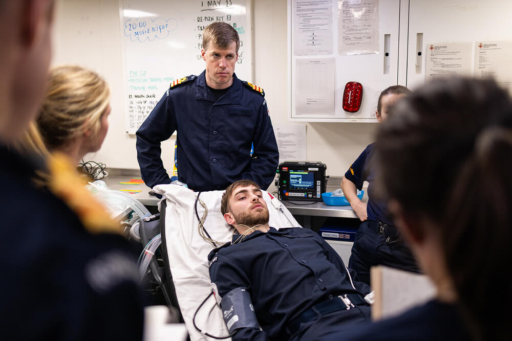 A colour image shows several Royal Navy medical officer students in a learning session. In the centre, a cadet lies back on an examination chair. He has a blood pressure band and several wires attached to him. They all wear blue Royal Navy work uniforms.