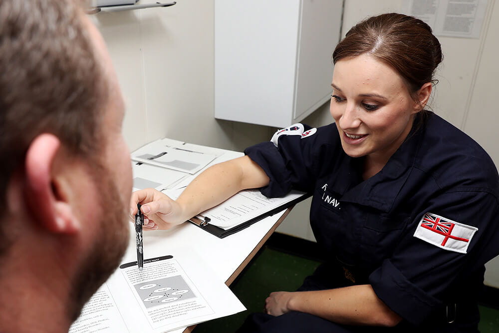 A colour images shows a Royal Navy nurse sitting at a desk with a clipboard and several printed sheets. She wears a blue Royal Navy work uniform. She is chatting with a patient and drawing attention to an informational diagram in one of her print-outs. The image is cropped and the patient's side profile is visible.
