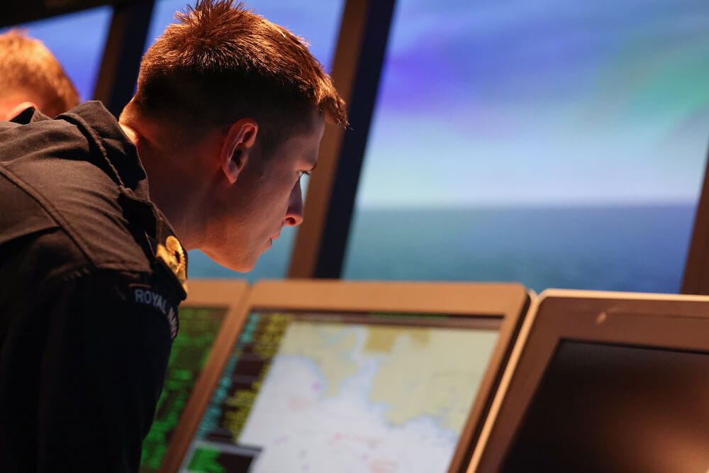 A colour image shows a Royal Navy meteorology officer examining geographic weather readings on a large digital screen. He wears a blue Royal Navy work uniform. In the background, the ocean can be seen through large windows on the ship's bridge.