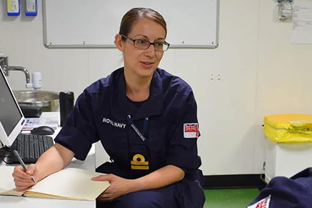 A colour photo shows a Royal Navy mental health nursing officer sitting at a desk and writing on a notepad. At the same time, she is chatting with a patient who is just out of frame; only a shoulder is visible. The officer wears a blue Royal Navy work uniform and glasses. In the background is a computer, a sink, and a dry-erase board.