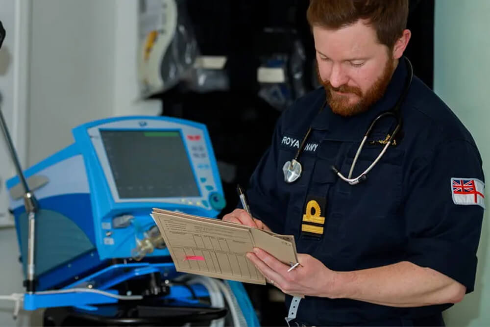 A colour image shows a Royal Navy nursing officer taking notes in a medical folder. He is wearing a blue Royal Navy work uniform and a stethoscope. In the background is a large and bright blue medical machine with a monitor and several buttons.