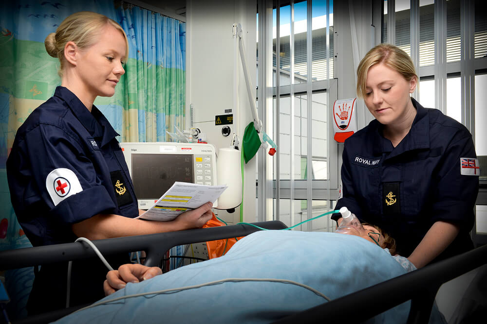 A colour image shows two Royal Navy medical servicemembers attending to a patient in a bed. They both wear blue Royal Navy work uniform. The patient lies flat on their back with a breathing appartus attached to their face. In the background are medical devices, monitors, and a privacy curtain.
