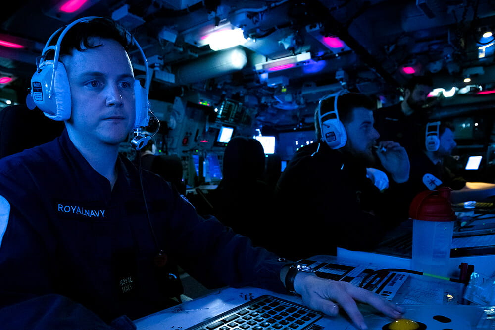 A colour image shows a Royal Navy intelligence specialist sitting at a workstation. He looks intently at equipment just out of frame. He is in a dark room illuminated by blue lights. In the background are three more intelligence specialists. Three of the specialists wear a protective headset. All four specialists wear a blue Royal Navy work uniform
