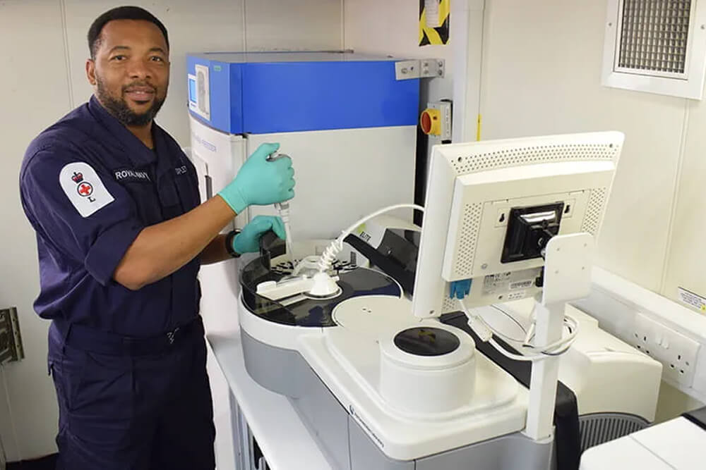 A colour image shows a Royal Navy biomedical scientist conducting lab work at a workstation with specialist equipment and a microscope. He wears a blue Royal Navy work uniform and sterile gloves.