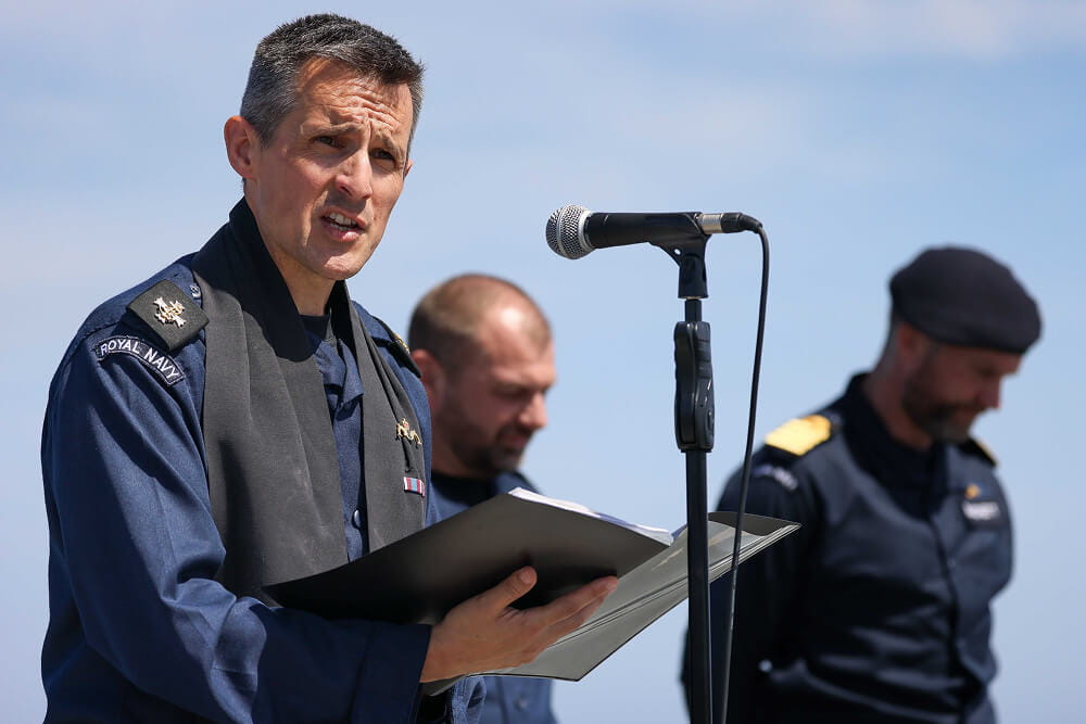 A colour image shows a Royal Navy chaplain standing at a microphone with a booklet in his hand. He wears a blue Royal Navy work uniform with a black cloth draped over his shoulders. In the background are two servicemen bowing their heads.