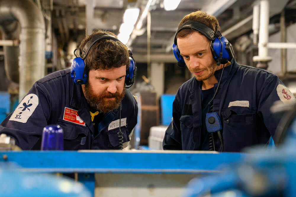 A colour image shows two Royal Navy engineers examining a part of a machine in a brightly lit room. They are both wearing blue Royal Navy work uniforms and protective headsets.