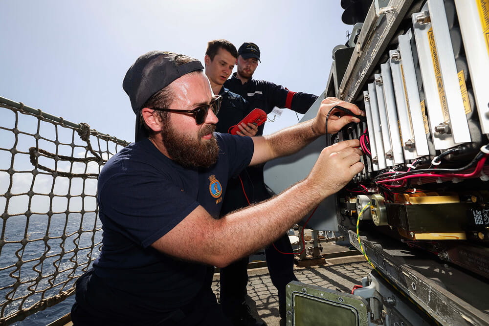 A colour image shows three Royal Navy engineers examining an electrical board on the deck of a ship. In the foreground, one technician handles specialist equipment. In the background, two technicians monitor readings on an external device. All three wear blue Royal Navy work uniforms.