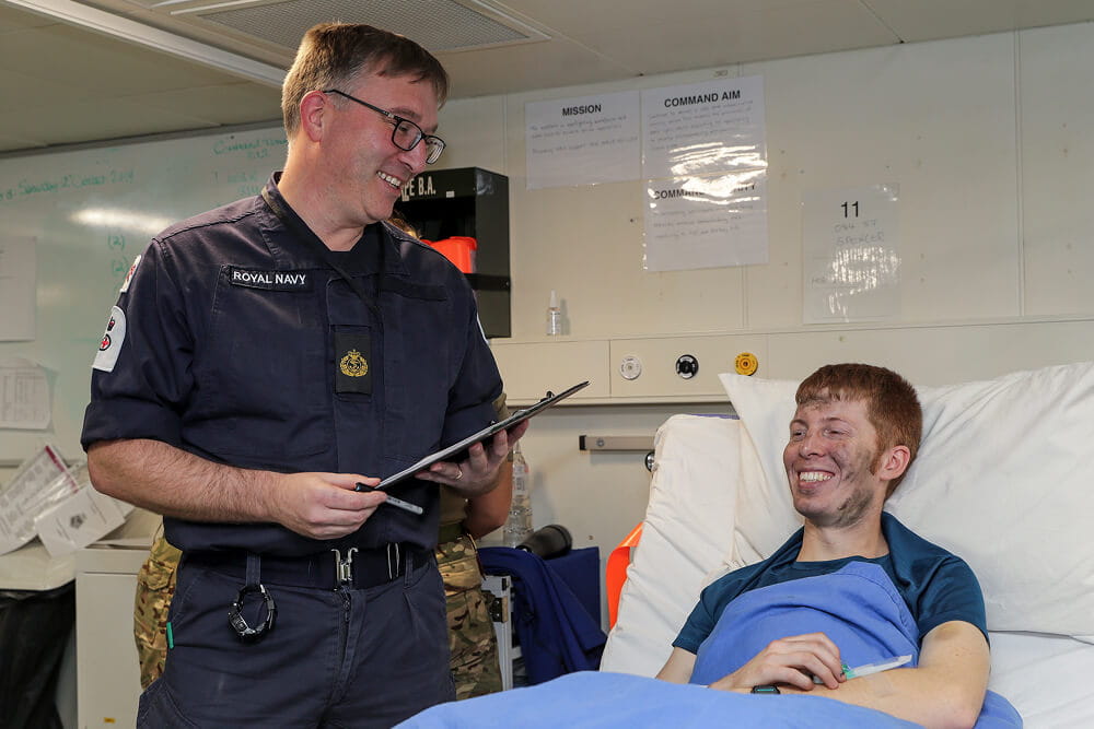 A colour image shows a Royal Navy nurse attending a patient's bedside. He wears a blue Royal Navy work uniform and holds a clipboard in his hands. The patient smiles while sitting upright in an infirmary bed.
