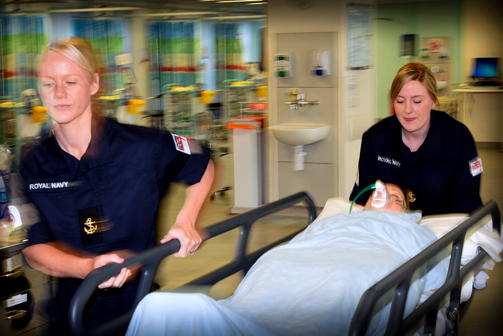 A colour image shows two Royal Navy medical students pushing a guerney through a bright infirmary. They both wear blue Royal Navy work uniforms with badges. A patient lies still on the guerney with a breathing apparatus around their face. The image is slightly blurred due to the rapid motion.