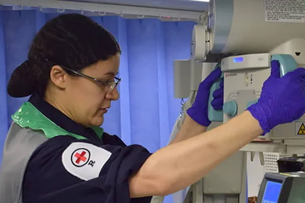 A colour photo shows a Royal Navy radiographer operating a specialist machine. She wears a blue Royal Navy work uniform, sterile gloves, a sterile vest, and glasses.