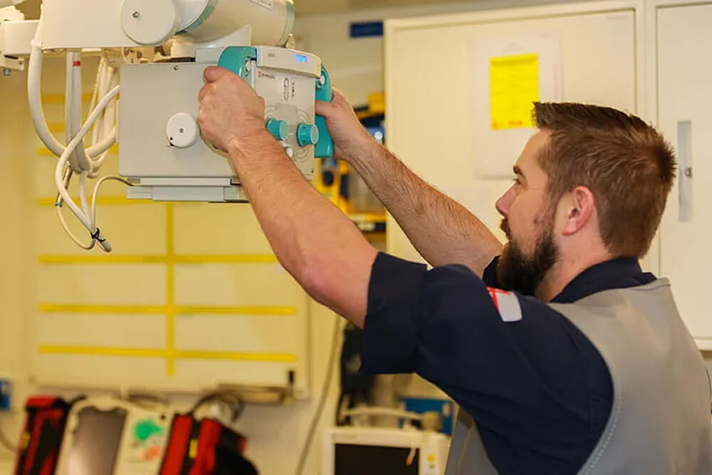 A colour image shows a Royal Navy radiographer handling specialist equipment. He wears a blue Royal Navy work uniform. In the background are various pieces of medical equipment and supplies, a dry-erase board, and storage cupboards.
