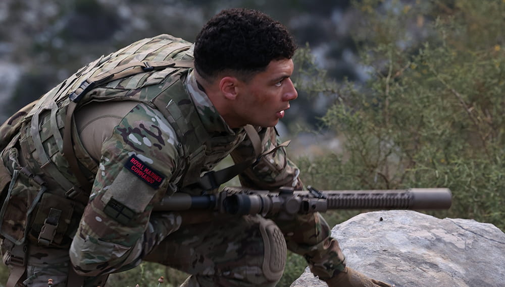 royal-navy-careers-life-in-the-royal-marines-02 Member of Royal Marines wearing full equipment and holding a weapon looking forward. Camouflaged amongst plants and rocks.