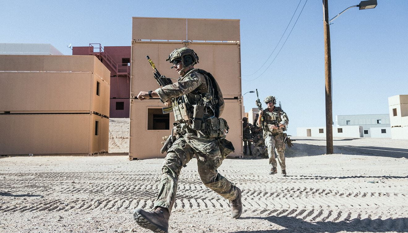 Two Royal Marines in the desert running across a road with buildings in the background.