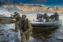 Two Royal Marines in camouflage on a coastline with one Royal Marine using a gun on a boat and another Royal Marine driving the boat.
