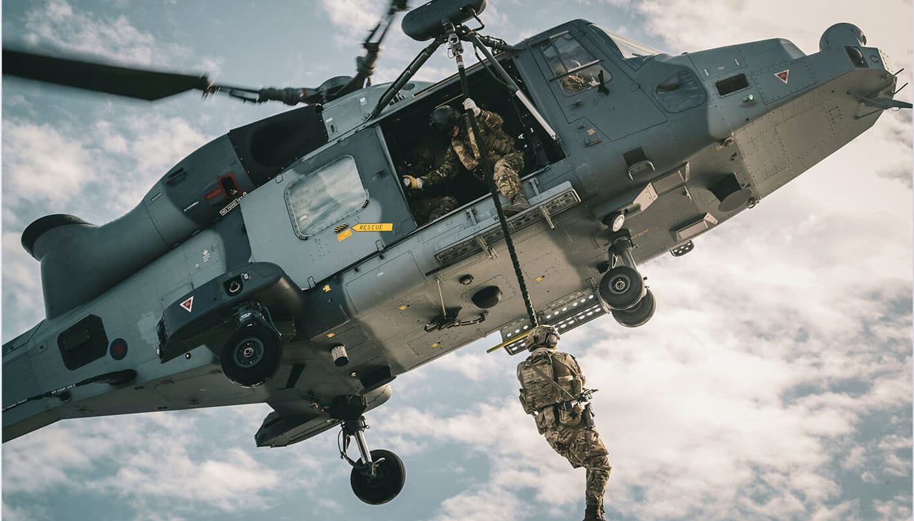 A Royal Marine is dangling from a helicopter on a cord with another Royal Marine assisting them.
