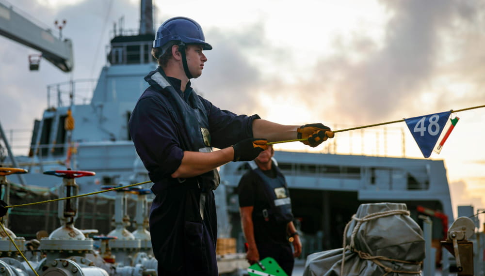 RFA crew member wearing a helmet and gloves pulls a rope on the deck of a naval support ship at sunset, with another crew member and ship equipment visible in the background.