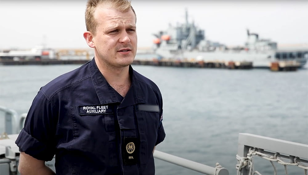 Navy personnel standing being interviewed with view of ship in the background. 