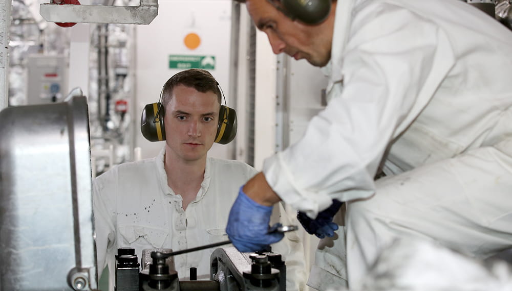 Two navy personnel work whilst wearing protective clothing and ear defenders. 
