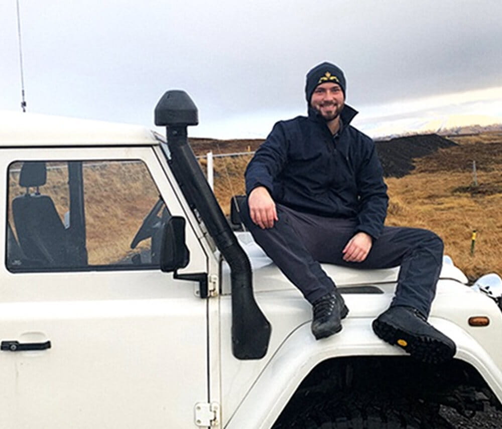 Submariner in uniform sitting on the bonnet of a Land Rover, smiling, with scenic countryside in background.