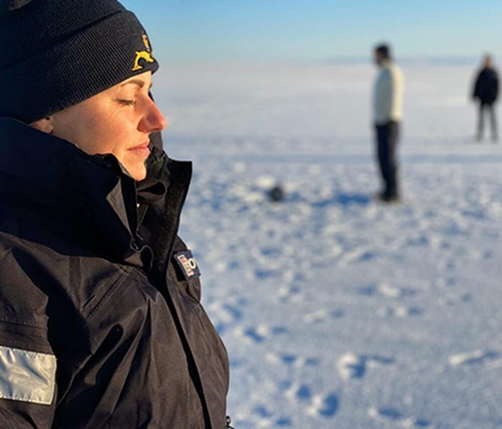 A submariner closing her eyes with sun on her face in the arctic while other crew member in the background are playing football