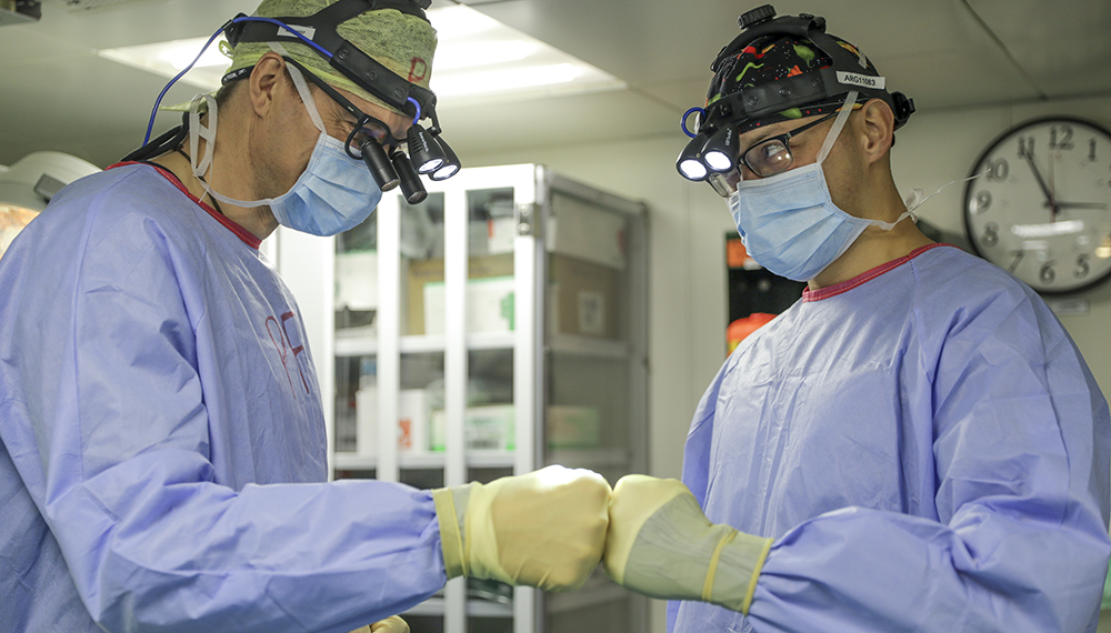 medics in light blue theatre robes with ocular headsets and yellow protective gloves fist bumping in clinical setting