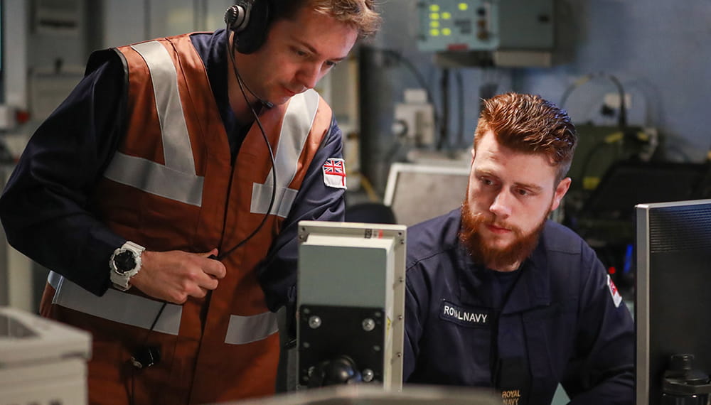 two uniformed crewmembers, one in orange high vis vest, of warfare Department in ship Operations Room