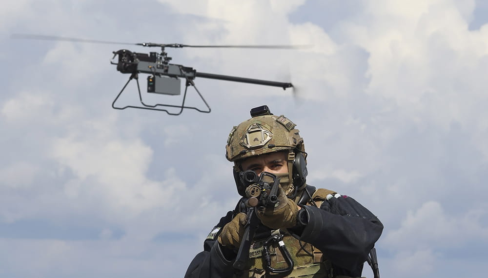Royal-Navy-Equipment-Commando-By-Air-07-Anduril-Ghost-MK4 An Anduril Ghost drone hovering over the head of a marine who is aiming a weapon at the camera