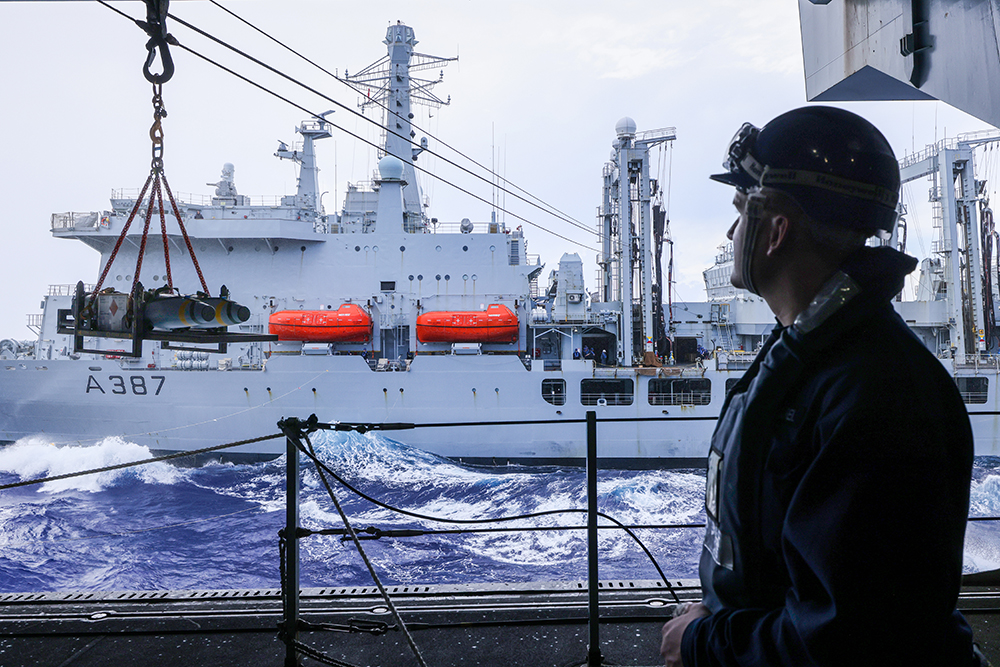 Royal Navy sailor in hardhat looks from the deck of one ship to another during a supply transfer