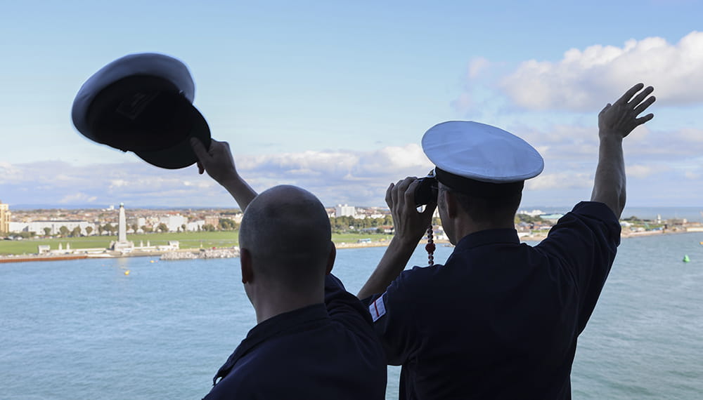 Two sailors on boat looking to shore with back to camera, one waving hat