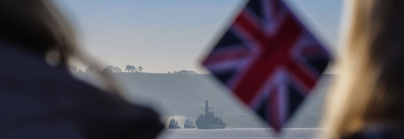 A British flag in the foreground with HMS Montrose coming into harbour in the background.