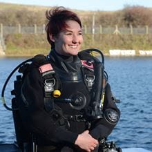 Female Navy diver in full gear smiles whilst looking off into the distance 