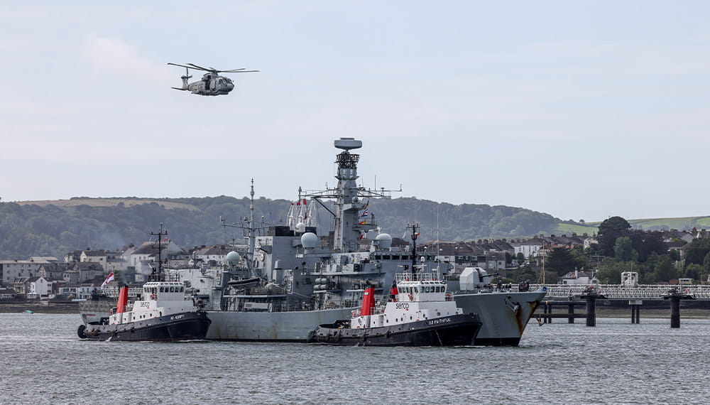Context image of a Type 23 frigate, 2 harbour patrol boats, and a helicopter flying above within the port of Plymouth