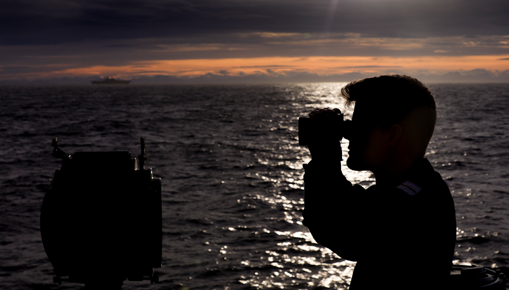 Navy sailor on a boat at sea glancing through binocular at dusk
